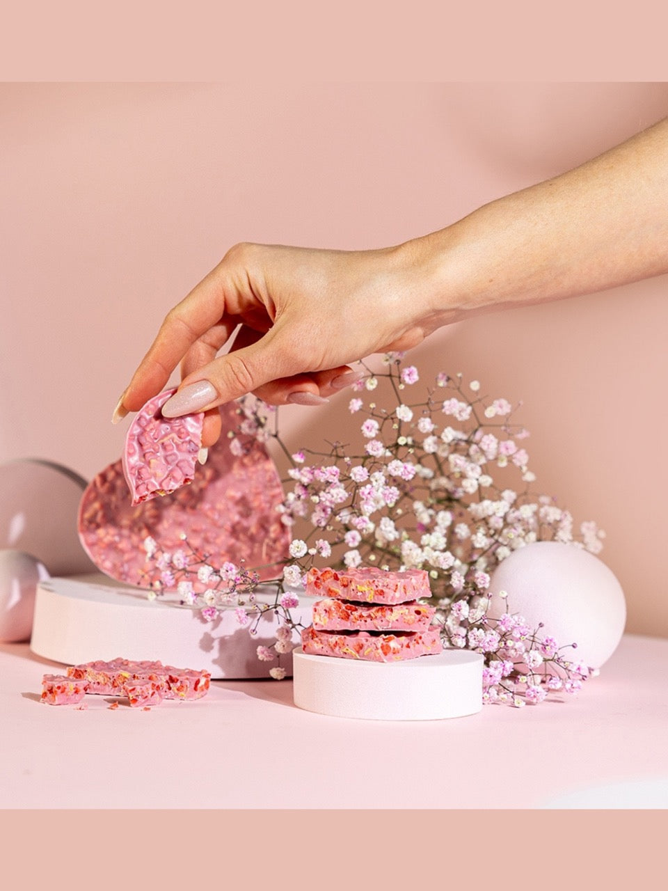 Detailaufnahme des Lauensteiner Erdbeer-Cornflakes-Herzens – rosa Schokoladenbruch mit Erdbeerstückchen und Cornflakes, dekorativ inszeniert mit Trockenblumen auf rosa Untergrund. Erhältlich in der Dreyer Confiserie Hameln.