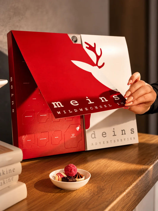 Geöffneter Meins & Deins Adventskalender von PETERS Pralinen mit aufklappbarer Vorderseite, Pralinentürchen und Schokoladenpralinen auf dem Tisch