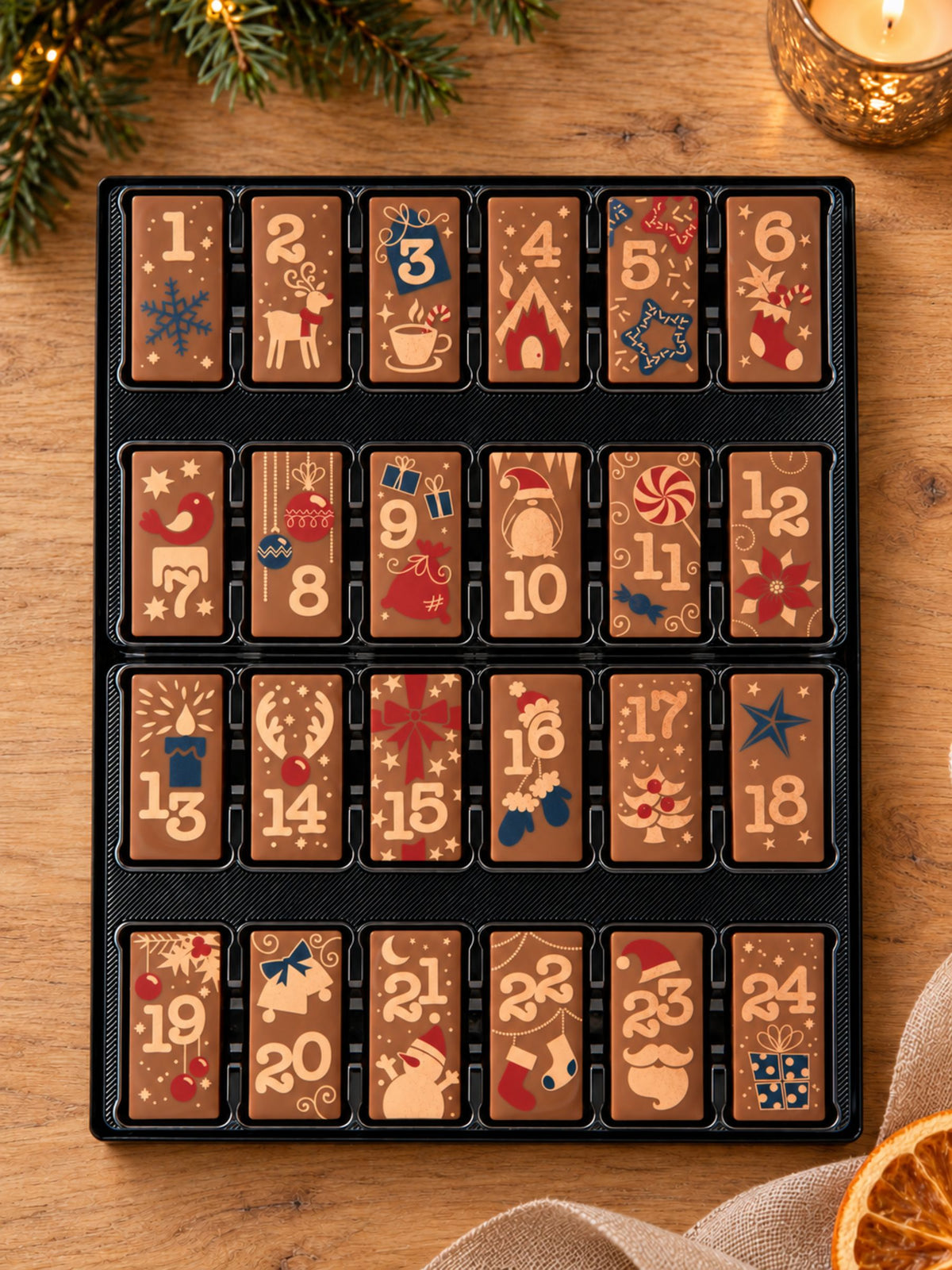 Weibler Adventskalender Domino Vollmilch 200g mit 24 nummerierten Schokoladendominos und weihnachtlichen Motiven auf einem Holztisch, dekoriert mit Tannenzweigen, Kerzenlicht und getrockneter Orangenscheibe