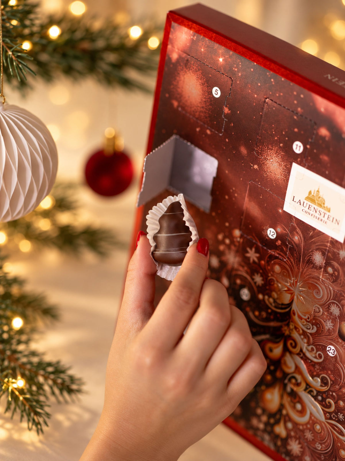 Lauensteiner Adventskalender Weihnachtsbaum 300g von Confiserie Lauenstein mit geöffnetem Türchen, handgefertigter Schokoladenpraline und festlichem Weihnachtsbaummotiv in Rot und Gold