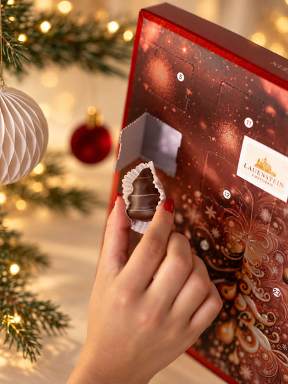 Lauensteiner Adventskalender Weihnachtsbaum 300g von Confiserie Lauenstein mit geöffnetem Türchen, handgefertigter Schokoladenpraline und festlichem Weihnachtsbaummotiv in Rot und Gold