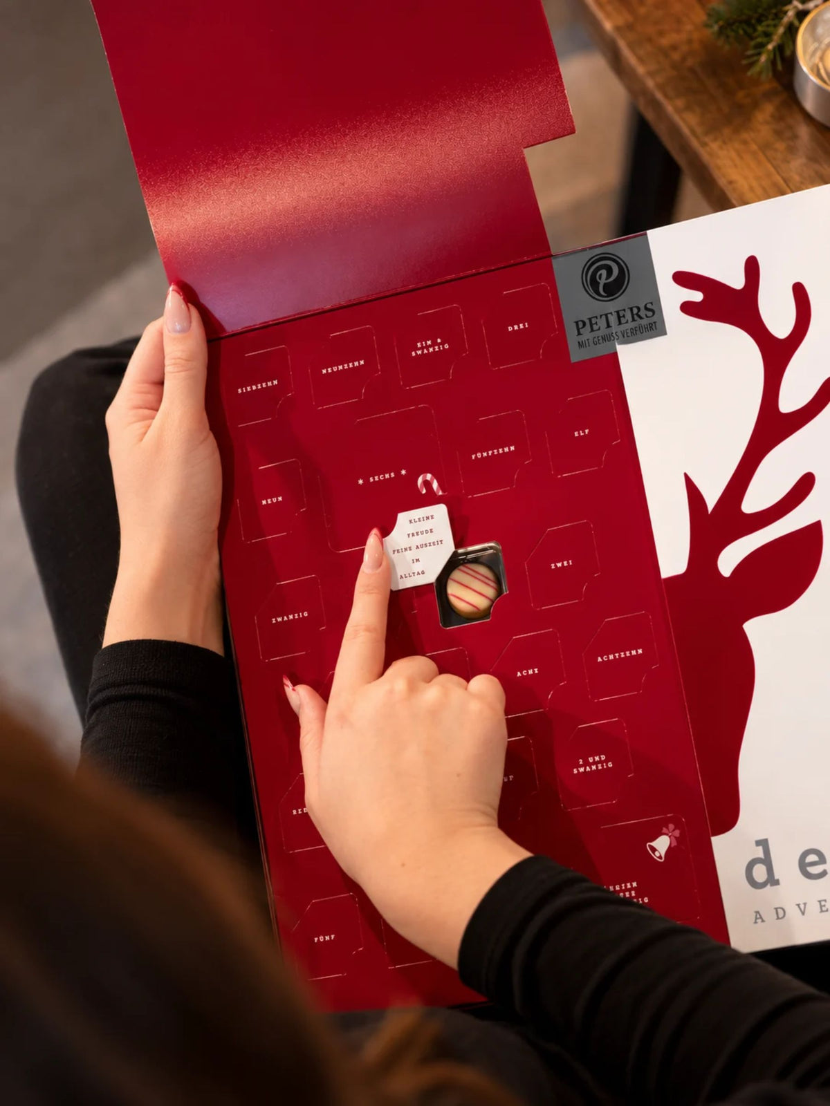 Geöffneter Meins & Deins Adventskalender von PETERS Pralinen mit aufklappbarer Vorderseite, Pralinentürchen und Schokoladenpralinen auf dem Tisch
