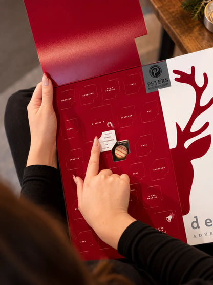 Geöffneter Meins & Deins Adventskalender von PETERS Pralinen mit aufklappbarer Vorderseite, Pralinentürchen und Schokoladenpralinen auf dem Tisch